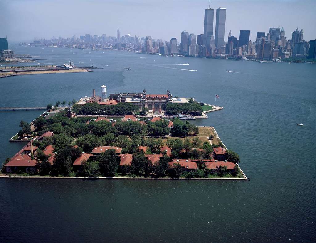 photo shows an aerial view of ellis island, with blue waters surrounding it, and the new york skyline behind.